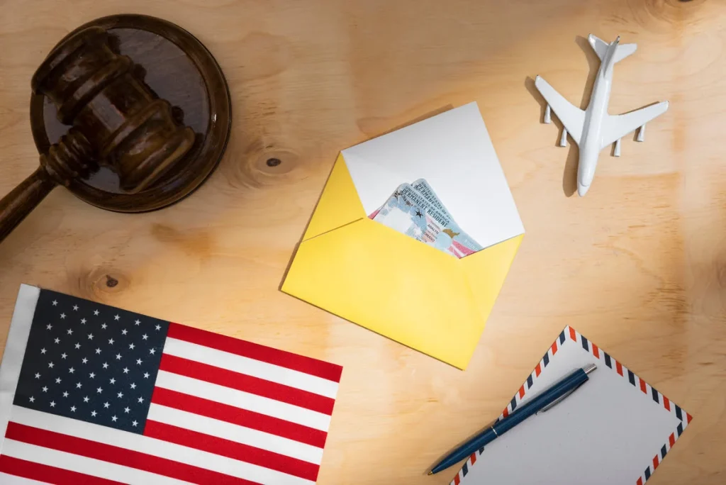 Top view of a Permanent Resident Card (Green Card) next to the US flag, a judge's gavel, and a toy airplane on a wooden table.