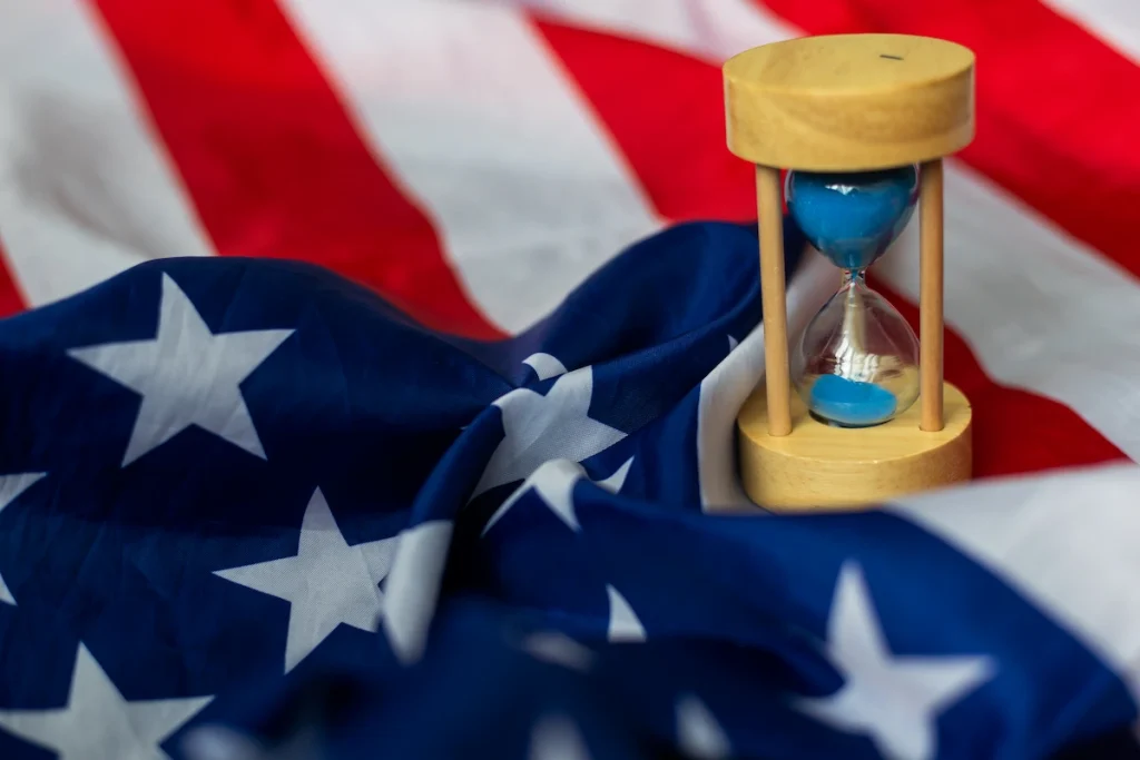 Reloj de arena frente a una bandera de Estados Unidos que representa los tiempos de procesamiento de la certificación laboral PERM