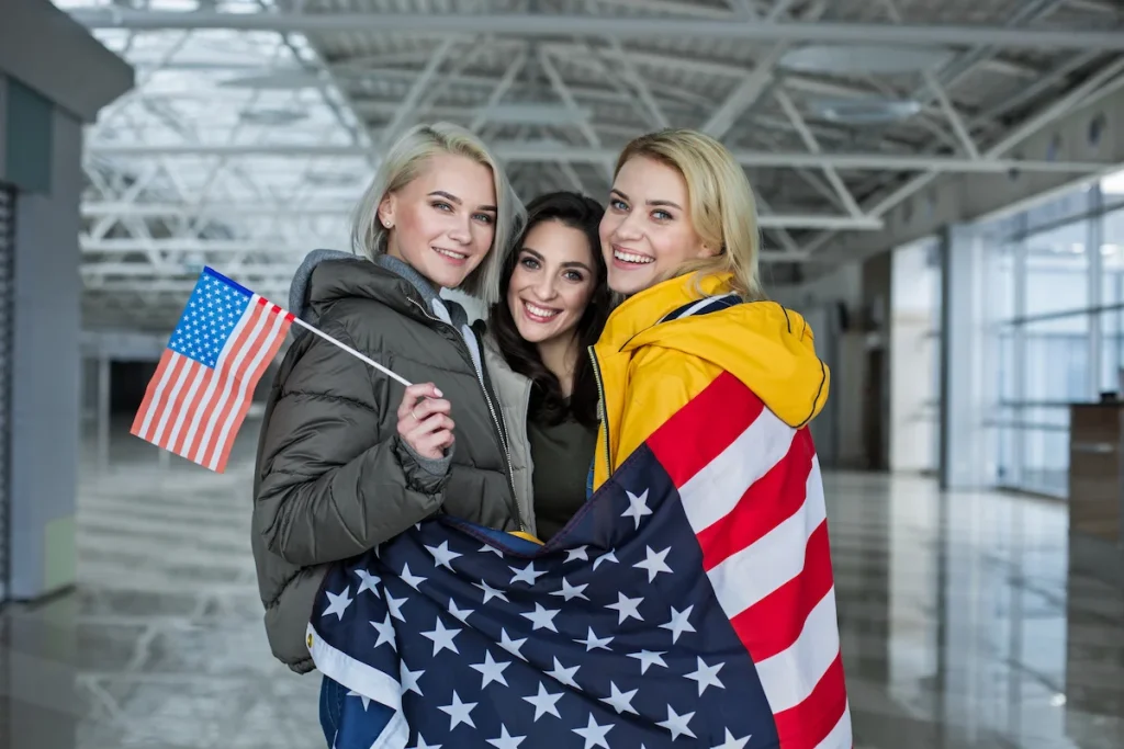 Three happy women wrapped in a U.S. flag celebrating employment avenues when asking can I sponsor a friend for a green card.